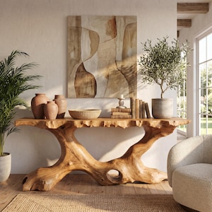 May include: A console table crafted from a single piece of light-toned natural wood with a live edge top and a sculptural, root-like base. The table holds three terracotta vases, a woven bowl, a stack of books, and a potted olive tree.