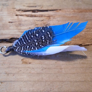 Puede incluir: Una pluma azul y blanca con manchas negras unida a un anillo plateado. La pluma es un elemento decorativo para un proyecto artesanal.