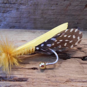 Puede incluir: Un señuelo de pesca con mosca amarillo y marrón con una cola de plumas y una cuenta dorada en el anzuelo.