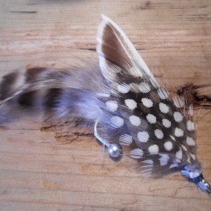 Puede incluir: Una pluma blanca y marrón manchada con una cuenta de plata y un gancho de plata. La pluma está sobre una superficie de madera.