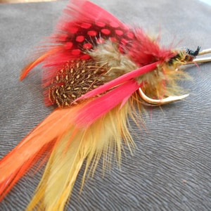 Puede incluir: Un señuelo de pesca con mosca de plumas rojas, naranjas y amarillas con un anzuelo dorado.