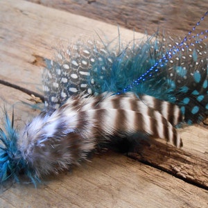 Puede incluir: Un arreglo de plumas decorativo con plumas azules, blancas y negras. Las plumas están dispuestas en forma de abanico y tienen un acento azul brillante.