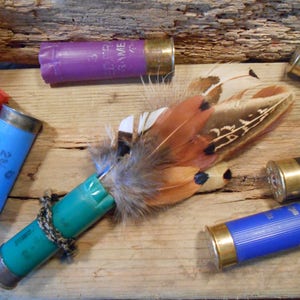 May include: A teal blue shotgun shell with a brown and white pheasant feather attached to it. The shell is surrounded by other shotgun shells in various colors, including purple, red, and blue.