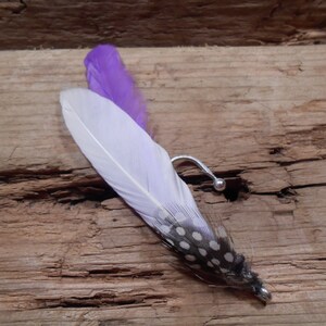 Puede incluir: Una pluma blanca con manchas negras y una punta morada unida a un gancho plateado.
