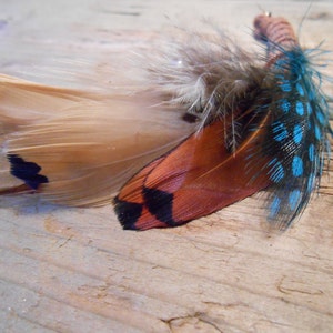 Puede incluir: Una pinza para el pelo de plumas con plumas marrones, negras y azules. Las plumas están dispuestas en un patrón decorativo.