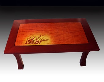May include: A rectangular wooden coffee table with a red wood frame and a light wood top. The top features a dark wood inlay of a stylized plant design.