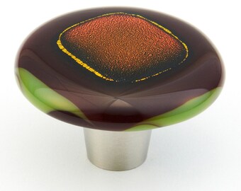May include: A round, brown glass cabinet knob with a green swirl design and a gold-rimmed, textured, orange square in the center. The knob has a silver metal base.
