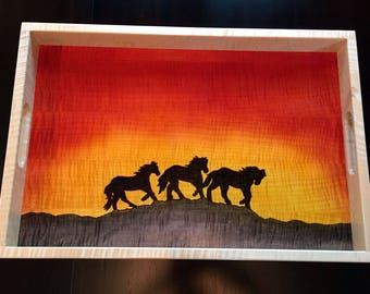 May include: A wooden serving tray with a sunset scene painted on the bottom. The sunset is a gradient of orange, yellow, and red. There are four black silhouettes of horses running across the bottom of the tray.
