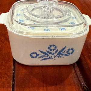 May include: A vintage white casserole dish with a clear lid and a blue floral design. The lid has a timer dial. The dish has a square shape with rounded corners and small handles on each side. A classic kitchenware item.