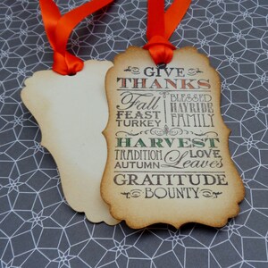 Puede incluir: Dos etiquetas decorativas de color crema con cintas naranjas. Una etiqueta presenta las palabras "GIVE THANKS", "FALL", "HARVEST" y otras palabras tem&aacute;ticas de oto&ntilde;o en un estilo vintage. Las etiquetas est&aacute;n sobre un fondo gris y blanco con patrones geom&eacute;tricos.