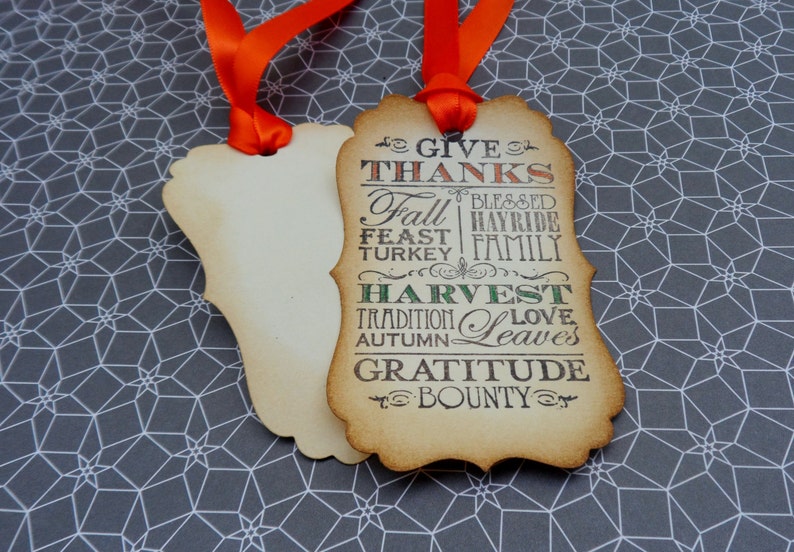 Puede incluir: Dos etiquetas decorativas de color crema con cintas naranjas. Una etiqueta presenta las palabras "GIVE THANKS", "FALL", "HARVEST" y otras palabras tem&aacute;ticas de oto&ntilde;o en un estilo vintage. Las etiquetas est&aacute;n sobre un fondo gris y blanco con patrones geom&eacute;tricos.