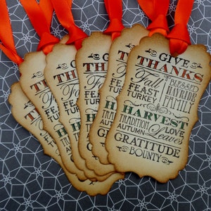 Puede incluir: Un conjunto de etiquetas de regalo con tem&aacute;tica de Acci&oacute;n de Gracias con cintas naranjas. Las etiquetas son de color beige con un borde decorativo y presentan las palabras "Give Thanks", "Harvest" y otras palabras oto&ntilde;ales.