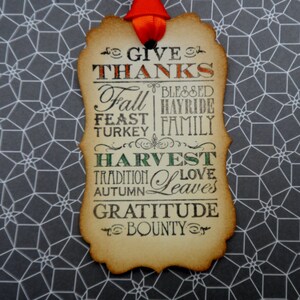 Puede incluir: Una etiqueta decorativa con borde festoneado y cinta roja. La etiqueta presenta las palabras "GIVE THANKS", "Fall", "Feast", "Turkey", "Harvest", "Tradition", "Autumn", "Gratitude" y "Bounty". El fondo es un patr&oacute;n geom&eacute;trico gris y blanco.