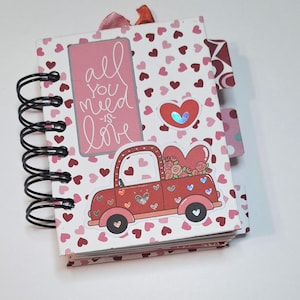 May include: A small pink and white notebook with a spiral binding. The cover features a red truck with a heart-shaped bed filled with flowers. The truck has a sticker of a heart with a blue and green iridescent finish. The cover also has a pink rectangle with the text "all you need is love" in white cursive writing. The background is covered in small pink and red hearts.