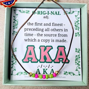 May include: A brown cord necklace with pink and green beads, displayed in a light green box. The card behind the necklace has the text "AKA" in pink letters and the words "MADE IN USA" on a red, white, and blue sticker.
