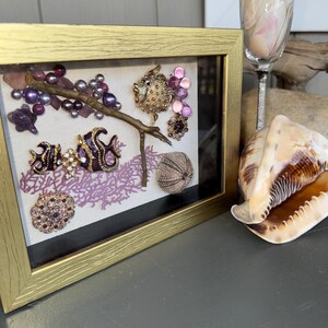 May include: A framed artwork featuring a gold frame, showcasing a collage of sea-themed elements. The artwork includes a branch with purple beads, decorative brooches, and a sea urchin. A seashell and a champagne flute are also visible.