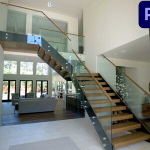 Op de afbeelding: Een modern huisinterieur met een glazen en houten trap die naar de tweede verdieping leidt. De trap heeft een glazen leuning en een houten handrail. Het huis heeft grote ramen en een wit interieur.