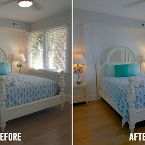 Op de afbeelding: Een wit bedframe met een blauw-wit gestreepte deken en twee turquoise kussens. Het bed staat in een slaapkamer met witte muren en houten vloer. De kamer heeft twee ramen met witte jaloezie&euml;n en twee lampen met palmboomvoeten.