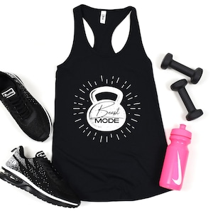 May include: Black tank top with a white graphic of a kettlebell and the text "Beast Mode". The tank top is lying flat on a white surface with a pair of black dumbbells, a black and white trainer, and a pink water bottle with a white Nike swoosh.