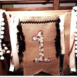 May include: A burlap banner with a large number one in the center. The number is decorated with a brown and white animal print. The banner is decorated with white pom poms, white and red ribbon, and brown burlap fringe.