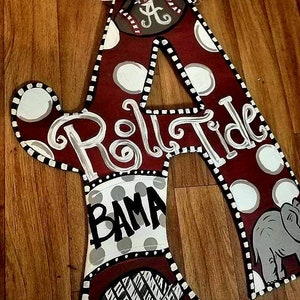 May include: A large wooden letter A painted in maroon with white polka dots and a black and white checkered border. The letter is decorated with the words "Roll Tide" and "BAMA" in white paint. The letter also features a gray elephant, a baseball with the letter A on it, and a gray and white striped pattern.