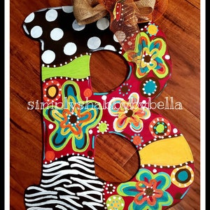 May include: A wooden letter B painted in a colorful, whimsical style. The letter is decorated with polka dots, stripes, flowers, and swirls in shades of red, yellow, green, blue, white, and black. The letter is accented with a burlap and orange mesh bow. The text "simply shabby by bella" is visible on the letter.