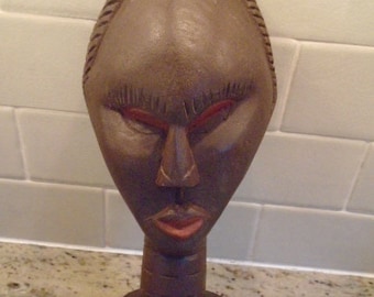 May include: A wooden sculpture of a person's head and neck. The sculpture is dark brown with red painted lips and eyes. The sculpture is carved with details on the face and neck.