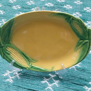 May include: An oval ceramic serving dish with a smooth yellow center and a raised, three-dimensional border featuring green corn husks and yellow corn kernels. The dish is supported by a clear plastic stand, showcasing the detailed botanical relief.