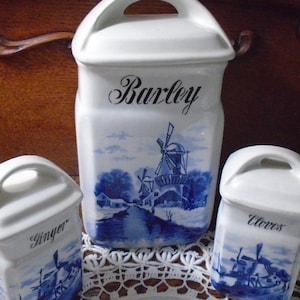 May include: Three white ceramic spice jars with blue windmill and landscape designs. The jars are labeled "Barley", "Ginger", and "Cloves". Each jar has a handle on top.