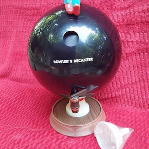 May include: A black bowling ball-shaped decanter with the text "BOWLER'S DECANTER" in white. A figurine of a man in a red shirt, blue trousers, and red shoes sits atop the decanter. The decanter has a wooden base and a spout.