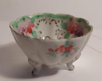 May include: A small, white porcelain bowl with three legs. The bowl is decorated with a floral pattern in shades of pink, red, green, and purple. The bowl has a scalloped edge.