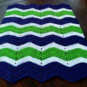 May include: A handmade crochet blanket with a chevron pattern. The blanket features alternating stripes of navy blue, white, and bright green. The blanket is made of soft yarn and is suitable for use as a throw or decorative piece.