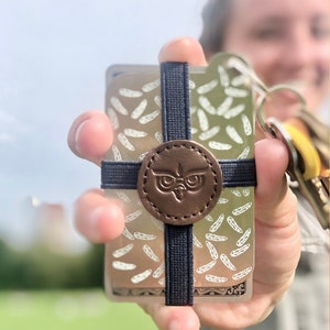 May include: A card holder with a brown leather emblem featuring an owl design. The cards have a feather pattern and are secured with a navy blue elastic band.
