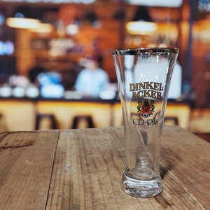 May include: A clear glass beer stein with a gold logo that says "Dinkel Acker CD-Pils". The logo features a crest with a lion and a crown.
