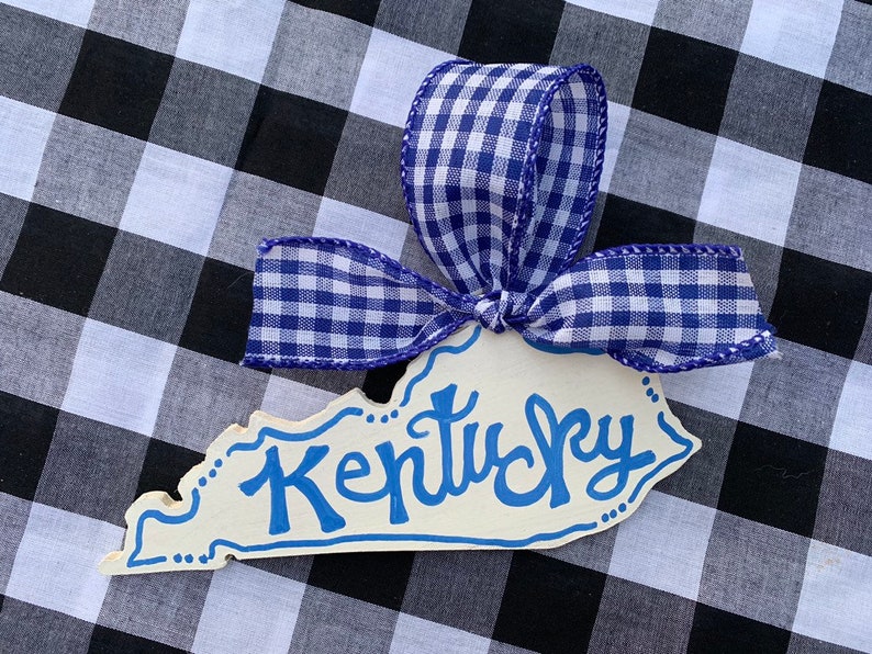 May include: A white wooden ornament shaped like the state of Kentucky with the word "Kentucky" written in blue paint. The ornament is decorated with a blue and white gingham ribbon.