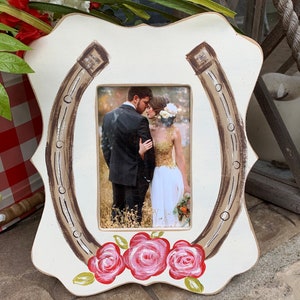 May include: White wooden picture frame shaped like a horseshoe with a brown painted horseshoe design. The frame has three red roses painted at the bottom and a photo of a couple inside the frame.