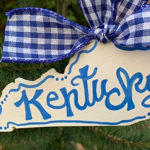 May include: A white wooden ornament shaped like the state of Kentucky with the word "Kentucky" written in blue paint. The ornament is decorated with blue and white gingham ribbon.