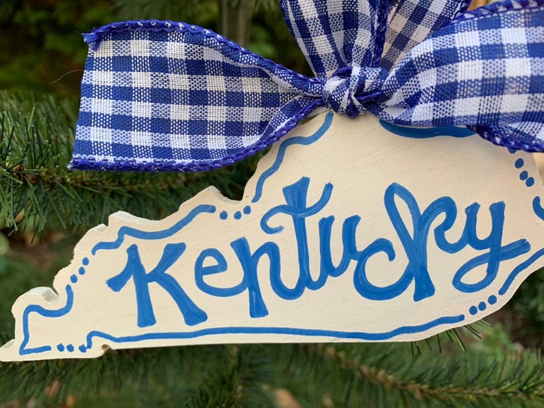 May include: A white wooden ornament shaped like the state of Kentucky with the word "Kentucky" written in blue paint. The ornament is decorated with blue and white gingham ribbon.