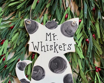 May include: A white wooden cat-shaped ornament with black painted spots and the text Mr. Whiskers in black lettering. The ornament features a silver wire hanger and a black and white gingham ribbon tied at the top for hanging display.