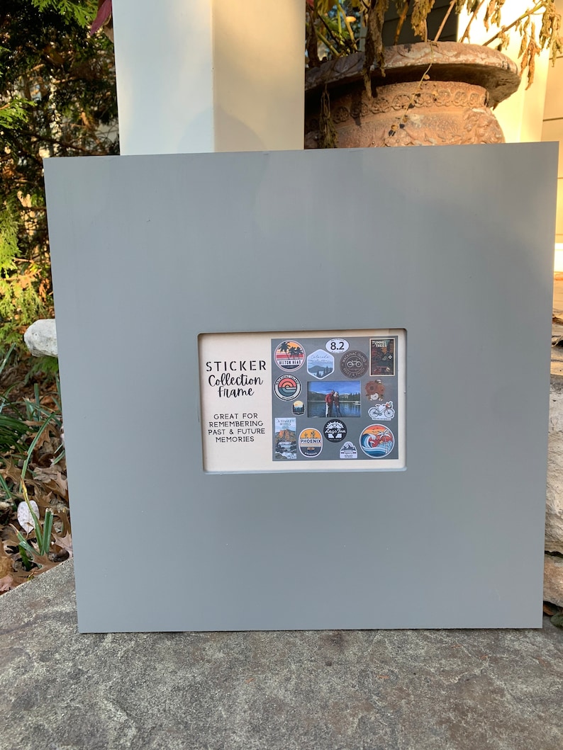 May include: Gray wooden frame with a white mat and a clear plastic insert. The insert has a collection of colorful stickers with outdoor themes. The text on the frame reads "Sticker Collection Frame" and "Great for remembering past & future memories".
