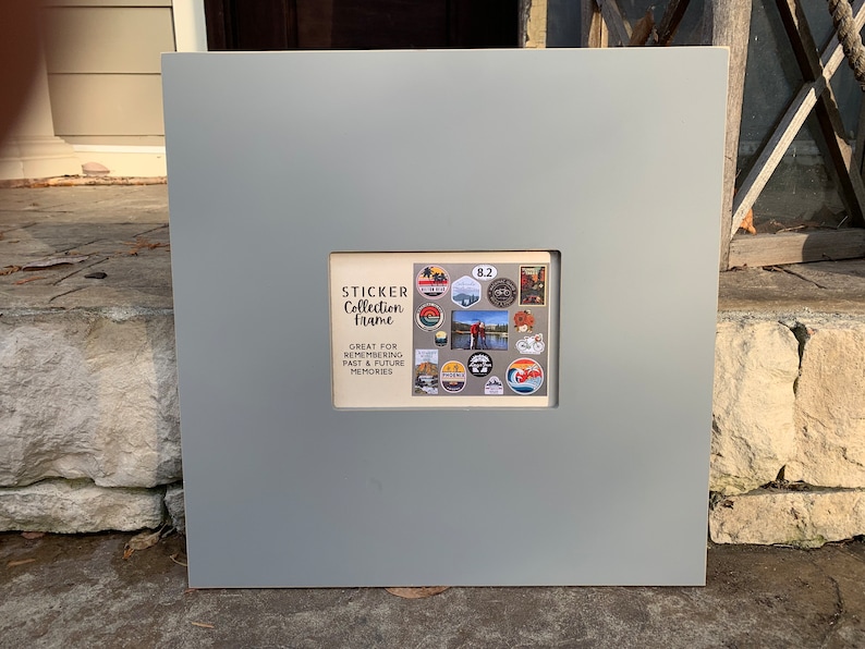 May include: Gray square frame with a smaller rectangular opening. The opening contains a collection of colorful stickers with a variety of designs. The text "STICKER Collection Frame" and "GREAT FOR REMEMBERING PAST & FUTURE MEMORIES" is printed on the frame.