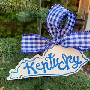 May include: A white wooden ornament shaped like the state of Kentucky with the word "Kentucky" written in blue paint. The ornament is decorated with blue and white gingham ribbon.