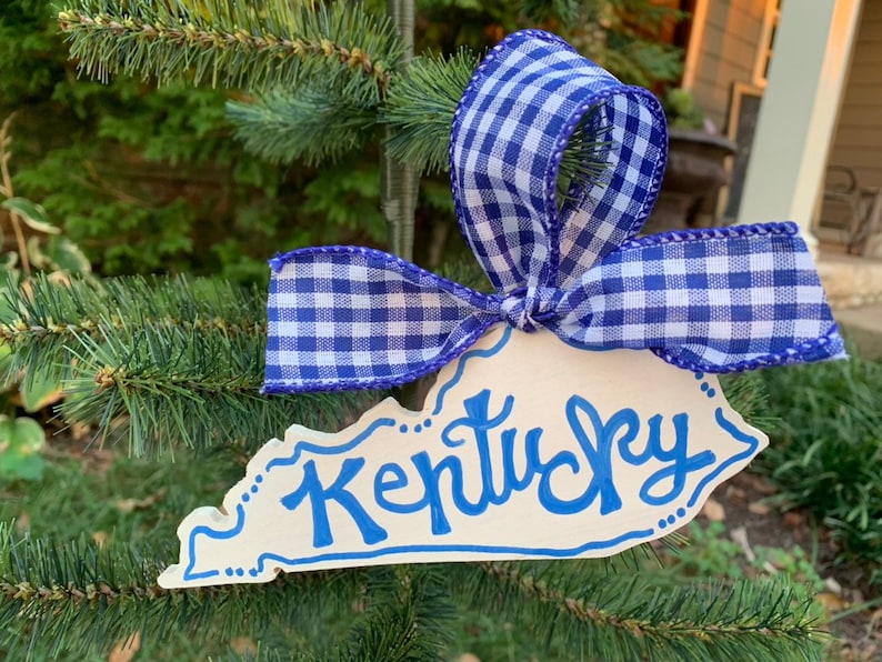 May include: A white wooden ornament shaped like the state of Kentucky with the word "Kentucky" written in blue paint. The ornament is decorated with blue and white gingham ribbon.