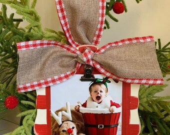 May include: A red and white checkered wooden ornament with a photo clip. The ornament is decorated with a brown burlap bow with red and white gingham trim. A photo of a child in a Santa suit and a dog wearing reindeer antlers is displayed in the clip.