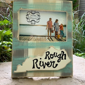 May include: A wooden picture frame with a light blue and gray plaid design. The frame has a photo of a family of three fishing on a lake. The text "I've Been Framed" is visible in the photo. The bottom of the frame has a cutout of the state of Kentucky with the text "Rough River" written in black.
