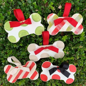 May include: Five wooden dog bone ornaments with different patterns and colors. The ornaments are decorated with red and white stripes, green and white polka dots, beige and white polka dots, red and white polka dots, and red and white stripes. Each ornament has a red ribbon tied around it.
