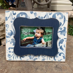May include: A white picture frame with a blue and white damask pattern. The frame has a dark blue inner border and a scalloped edge. The frame is holding a photo of a young girl with the text "We are Framed" in the photo.