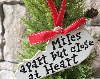 May include: White wooden ornament shaped like the state of Kentucky with the text "Miles apart but close at heart" in black lettering. The ornament is decorated with a red ribbon.