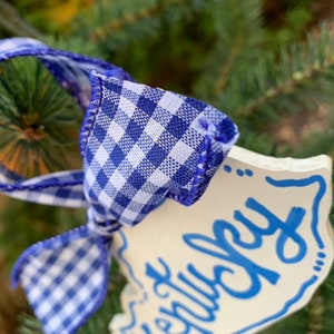 May include: White ceramic ornament with blue lettering that says "Merry Christmas". The ornament is tied with a blue and white gingham ribbon.