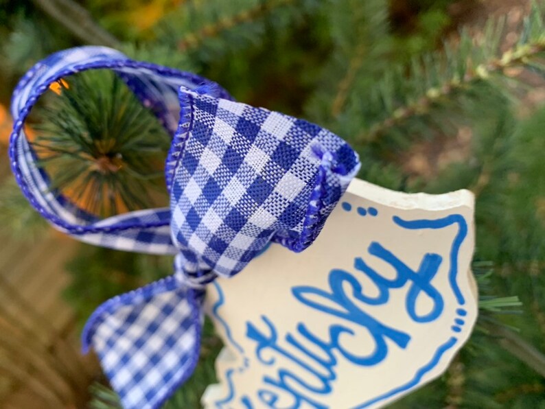 May include: White ceramic ornament with blue lettering that says "Merry Christmas". The ornament is tied with a blue and white gingham ribbon.